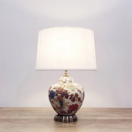 A round ceramic table lamp with a cream base with colourful floral designs and a metallic base