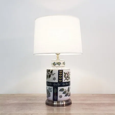 A cylindrical ceramic table lamp with white background and simple floral designs and a metal base