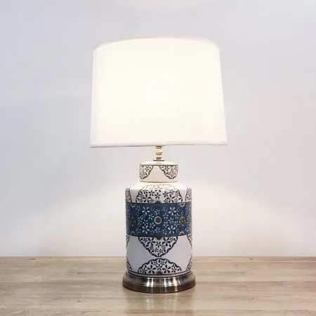 A cylindrical ceramic table lamp with blue, white & floral patterns against a white background and a metal base