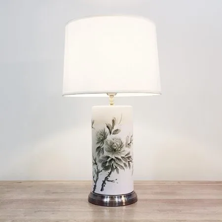 A tall cylindrical ceramic table lamp with ink-brush style grey floral painting against a white background and a metal base
