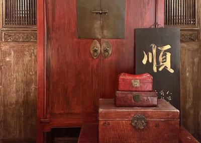 Chinese antique red wedding cabinet with square brass. This is surrounded by vintage leather boxes, and the background is a set of tall chinese antique doors