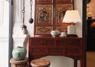 A small Zhejiang red lacquer 2-drawer table with carved floral drawer fronts and original butterfly-shaped brass hardware, displayed with a pair of richly carved Fujian cabinet doors used as a wall hanging, featuring original paintings in gold, teal, red and black. A pair of Fujian round stools with curved seat tops is visible to the left.