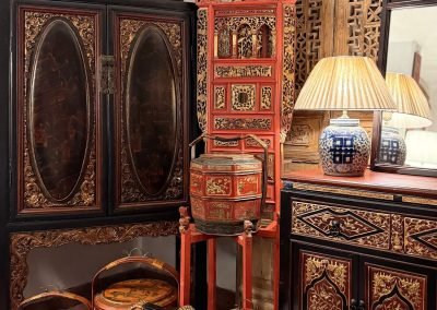 A black lacquered Chaozhou book cabinet with gold gilt carved bulbous door panels and carved skirting, displayed alongside a red and gold Fujian washbasin stand and a black side cabinet with red and gold carved panels. In front and on top are vintage lacquered tiered baskets from Wenzhou in various shapes, sizes and hand-painted designs.